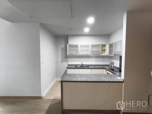 Condominium for rent in Khlong Toei, Bangkok - U-shaped kitchen with granite countertops, sink, cooktop, and glass-front wall cabinets.