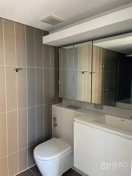 Condominium for rent in Khlong Toei, Bangkok - Second bathroom with vertical tile walls, vanity and toilet under a wide mirror.