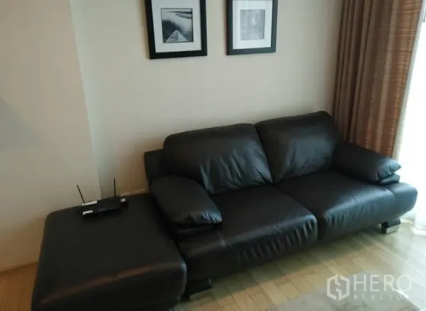 Condominium for rent in Khlong Toei, Bangkok - Living area featuring a black leather sofa, ottoman, framed wall art and full-height curtains.
