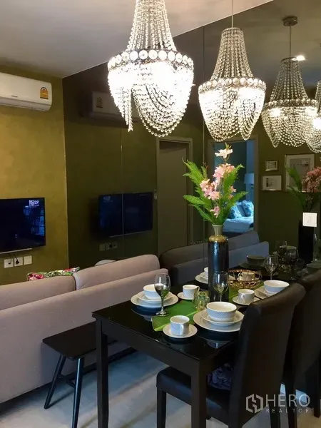 Condominium for rent in Khlong Toei, Bangkok - Dining table set for four beneath crystal pendant lights with mirrored wall and sofa beyond.