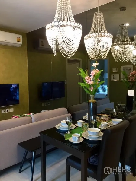 Condominium for rent in Khlong Toei, Bangkok - Dining table set for four beneath crystal pendant lights with mirrored wall and sofa beyond.