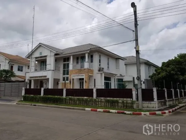 Detached House for sale in Lat Krabang, Bangkok - Corner-plot view of a modern white two-storey detached house with perimeter fence, hedges, balconies and tiled roof on a quiet residential street in Lat Krabang.