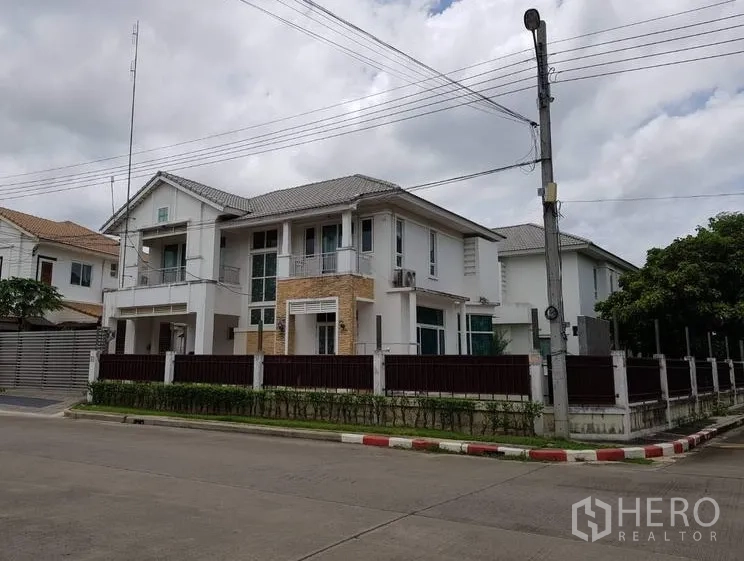 Detached House for sale in Lat Krabang, Bangkok - Corner-plot view of a modern white two-storey detached house with perimeter fence, hedges, balconies and tiled roof on a quiet residential street in Lat Krabang.