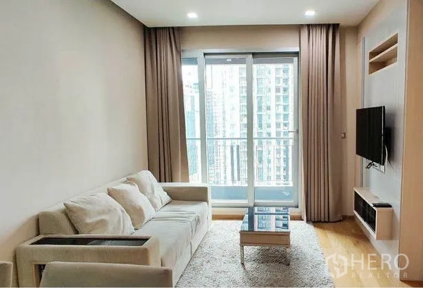 Condominium for rent in Ratchathewi, Bangkok - Bright condo living room with beige sofa, wall‑mounted TV, floor‑to‑ceiling windows and balcony city view.