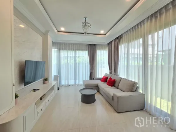独立式住宅 出租在Prawet, Bangkok - 采光充足的客厅，转角沙发、落地纱帘与内置电视墙。
