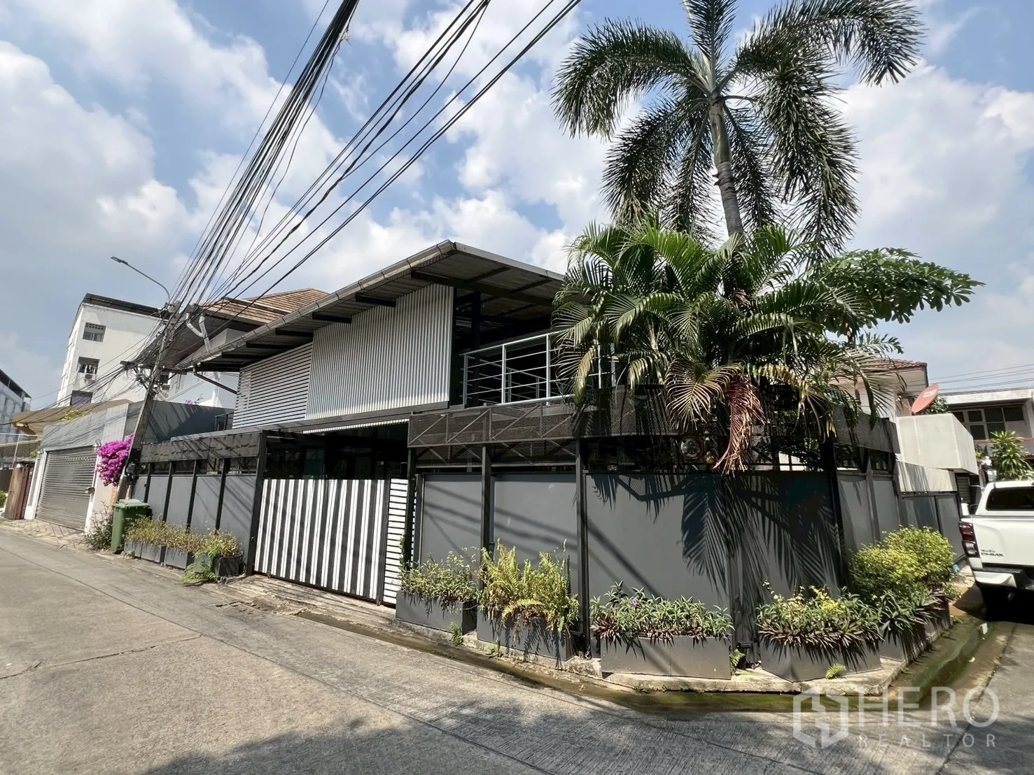Detached House for rent or sale in Wang Thonglang, Bangkok - Corner‑lot exterior with balcony, palm landscaping and sliding gate.