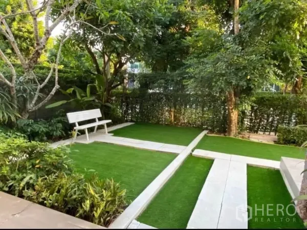 Detached House for rent or sale in Prawet, Bangkok - Corner garden with hedges, trees and a white bench set on synthetic lawn.