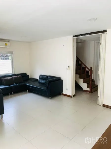 Townhouse for rent or sale in Huai Khwang, Bangkok - Bright living room with white tile floors, black leather sectional sofa and wooden staircase