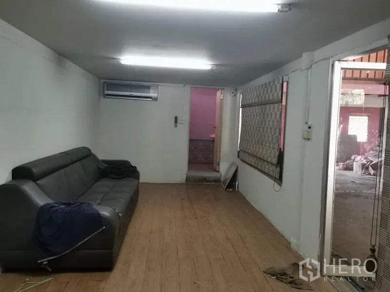 Warehouse for rent in Lam Luk Ka, Pathum Thani - Air-conditioned office room with a black sofa, wood-look flooring, fluorescent lights, and window blinds.