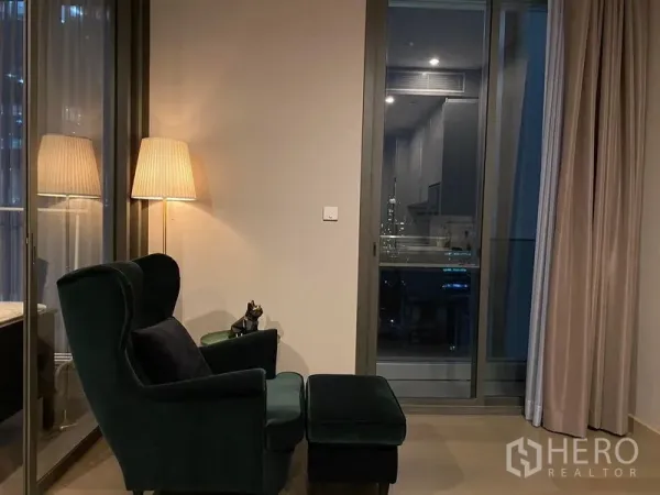 公寓 出租在Huai Khwang, Bangkok - 温馨阅读角，扶手椅与脚凳，旁有落地灯与阳台推拉门的夜景。