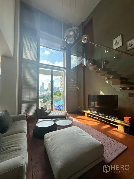 Condominium for rent or sale in Sathon, Bangkok - Double-height living room with sofa set, TV, and glass-railed staircase beside tall windows.