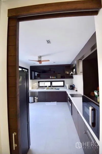 Townhouse for rent in Din Daeng, Bangkok - View into a long enclosed kitchen with white countertops, dark cabinets, ceiling fan and appliances.