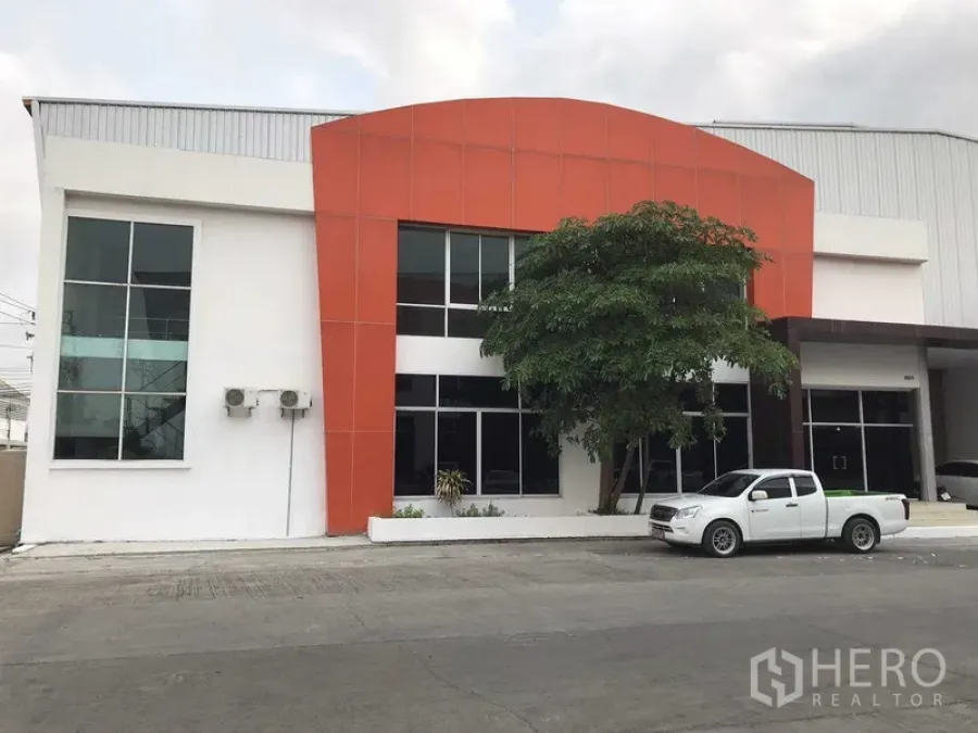 Factory for rent in Samut Prakan - Modern factory exterior with orange facade and two‑storey glass‑front office beside a driveway.