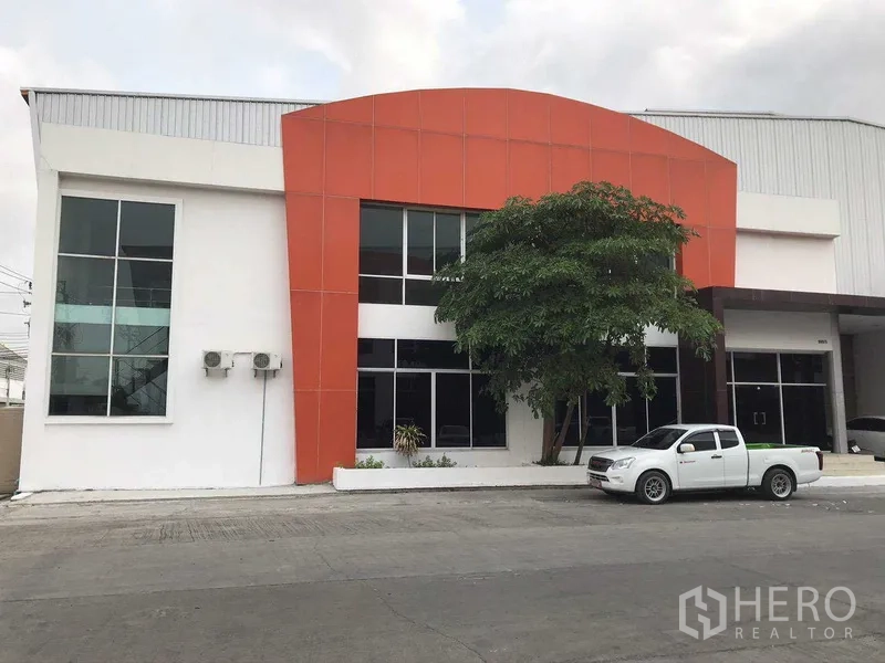 Factory for rent in Samut Prakan - Modern factory exterior with orange facade and two‑storey glass‑front office beside a driveway.