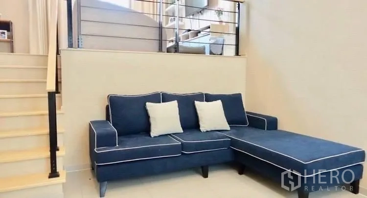 Townhouse for rent or sale in Suan Luang, Bangkok - Split-level living area featuring a blue L-shaped sofa and light wood stairs.