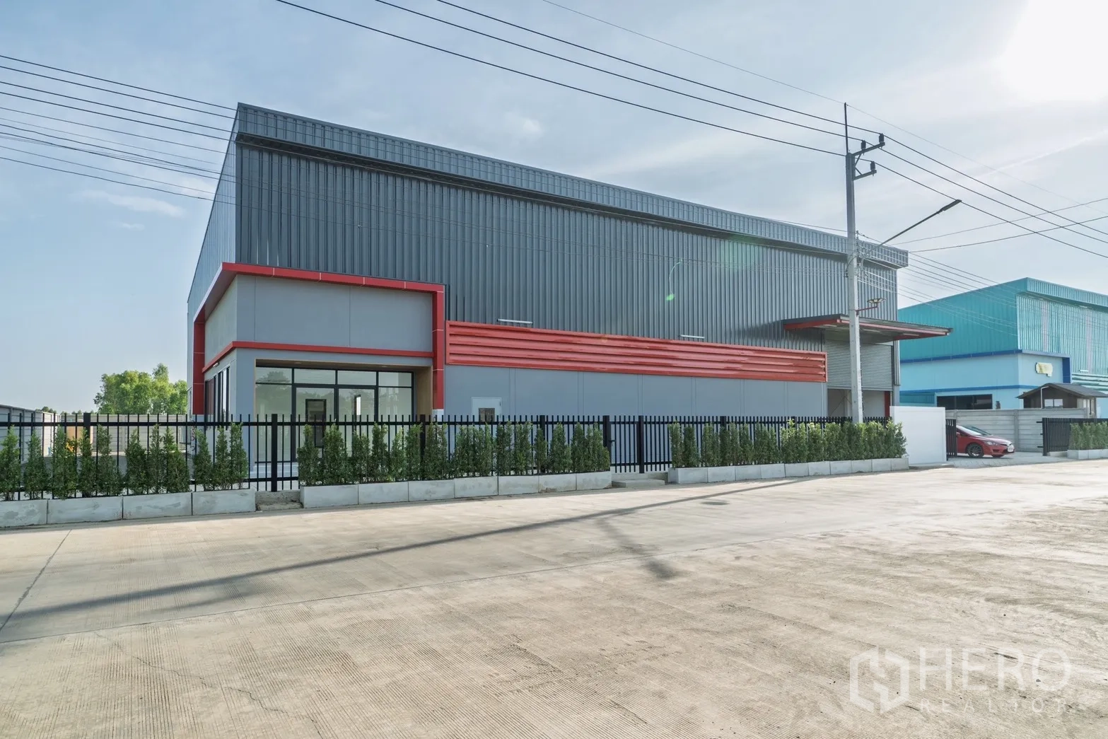 Factory for rent or sale in Lam Luk Ka, Pathum Thani - Modern factory exterior with grey metal cladding, red trim, gated fence and wide driveway.