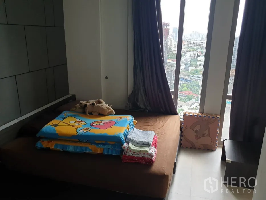 Condominium for rent in Khlong Toei, Bangkok - Bedroom with large windows, sweeping city view, dark blackout curtains, and a low platform bed.