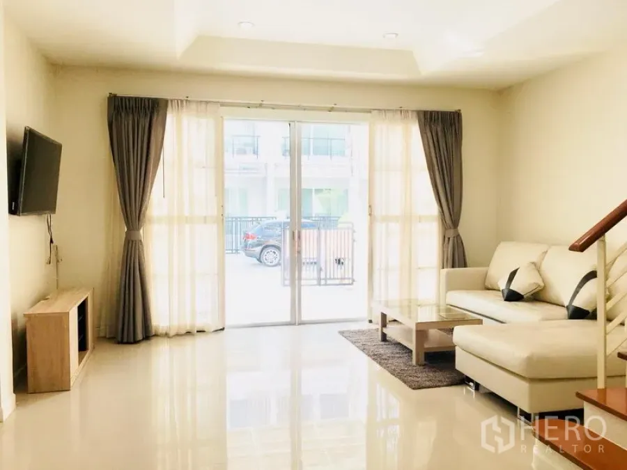 联排别墅 出租在Prawet, Bangkok - 采光充足的客厅，L形沙发、墙面电视与落地推拉玻璃门，连接前院与车位。