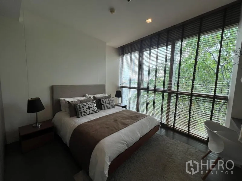 Condominium for sale in Khlong San, Bangkok - Bright bedroom with queen bed, bedside lamps, and tree-lined views through large windows.