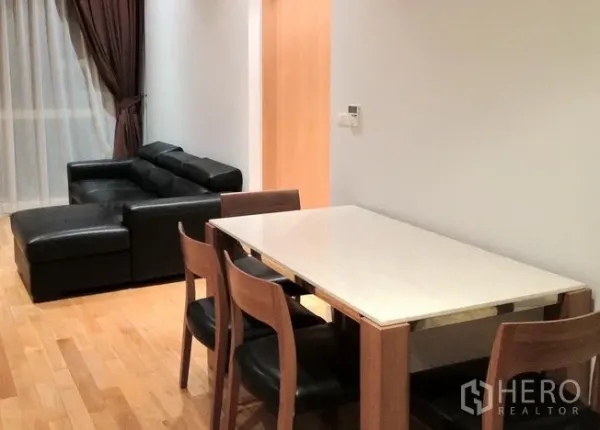 Condominium for rent in Khlong Toei, Bangkok - Dining area with a four-seat table beside a black leather sofa and a large window with sheer and blackout drapes.