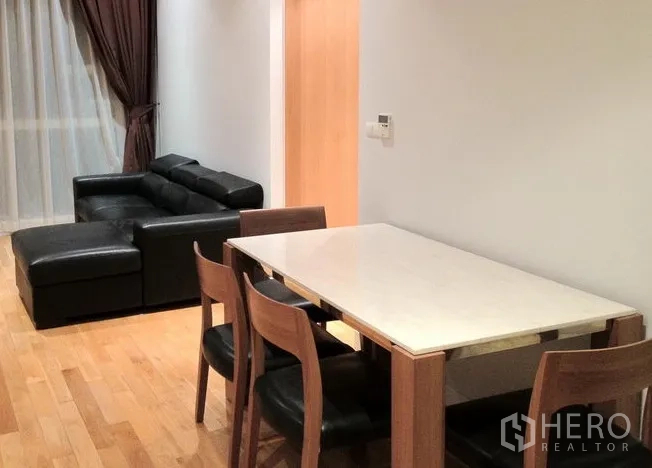 Condominium for rent in Khlong Toei, Bangkok - Dining area with a four-seat table beside a black leather sofa and a large window with sheer and blackout drapes.