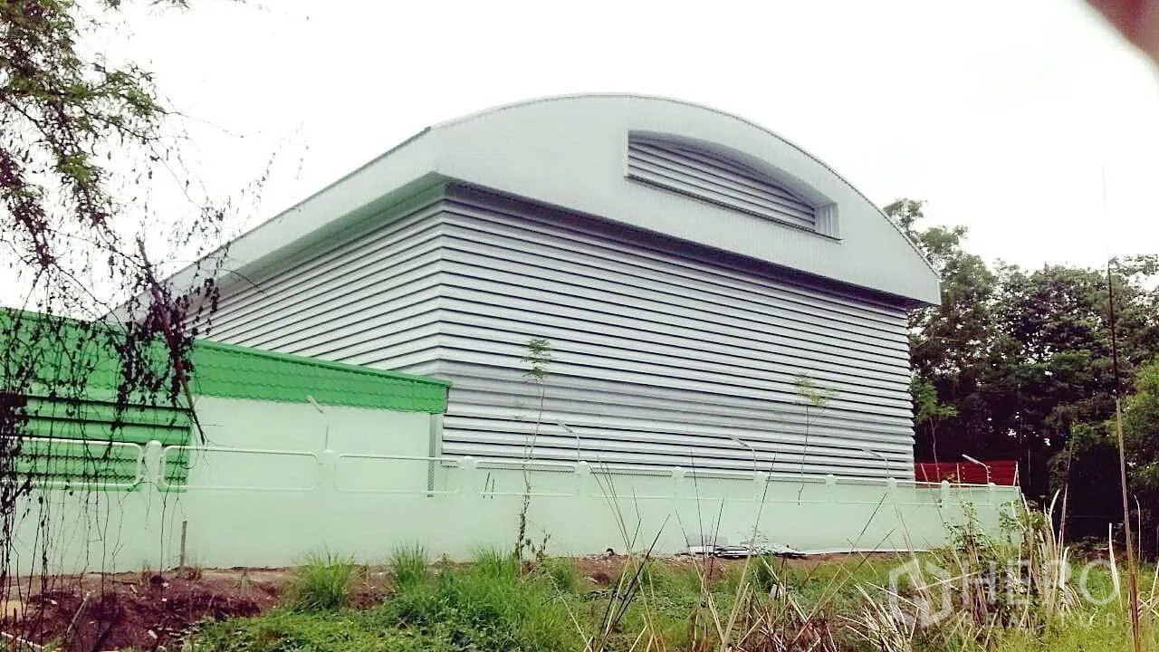 Factory for sale in Pathum Thani - Rear view of corrugated metal warehouse with curved roof and boundary wall amid greenery.