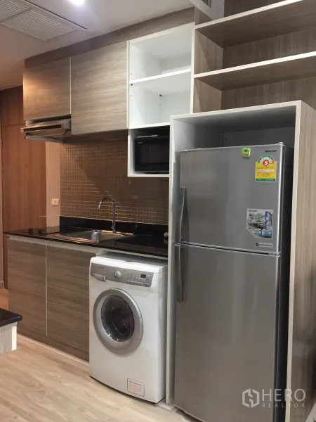 Condominium for rent or sale in Bangkok - Compact kitchenette with fridge, washing machine, sink and wood cabinets.
