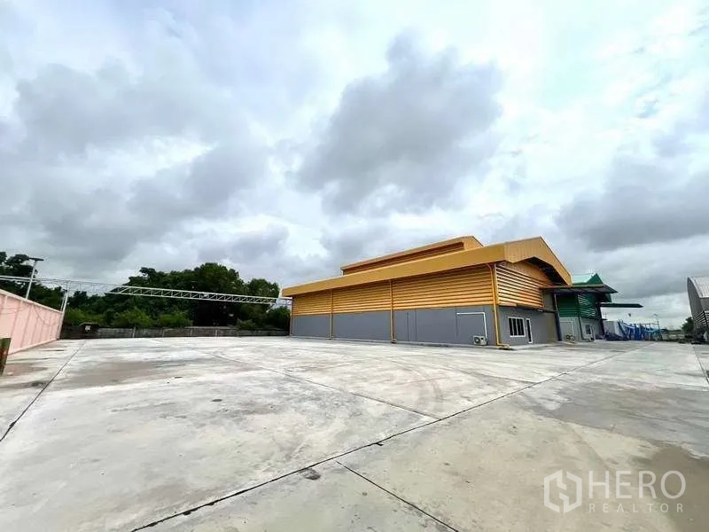 คลังสินค้า ให้เช่าในLat Lum Kaeo, Pathum Thani - ลานคอนกรีตกว้างและอาคารโกดังโครงเหล็กสีเหลืองภายนอกท่ามกลางท้องฟ้ามีเมฆ.