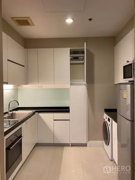 Condominium for rent in Khlong Toei, Bangkok - Modern L-shaped kitchen with white cabinets, built-in oven, fridge and washing machine.
