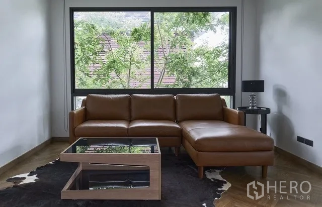 Townhouse for sale in Watthana, Bangkok - Upstairs lounge with a brown L-shaped sofa by a large window.