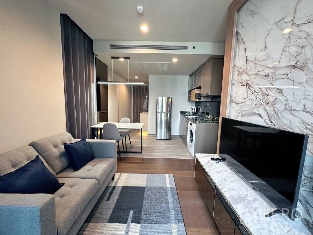 คอนโด for rent in Khlong Toei, Bangkok - ห้องนั่งเล่นและรับประทานอาหารสไตล์โมเดิร์น มีโซฟาสีเทา ผนังทีวีลายหินอ่อน ทีวีจอแบน ครัวเปิดพร้อมตู้เย็นและเครื่องซักผ้า และโต๊ะกินข้าวพร้อมโคมไฟแขวน