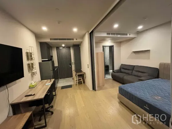 Condominium for rent in Watthana, Bangkok - Studio-style layout showing bed, sofa, entryway, kitchenette and wood floors.