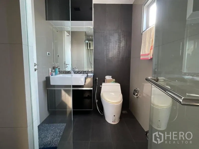 Condominium for sale in Khlong Toei, Bangkok - Second bathroom with glass shower, vanity and toilet in dark tile scheme.
