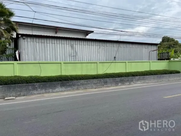 คลังสินค้า ขายในLam Luk Ka, Pathum Thani - ตัวโกดังผนังเมทัลชีทยาวอยู่หลังรั้วสีเขียวริมถนนสองเลนที่เงียบสงบ