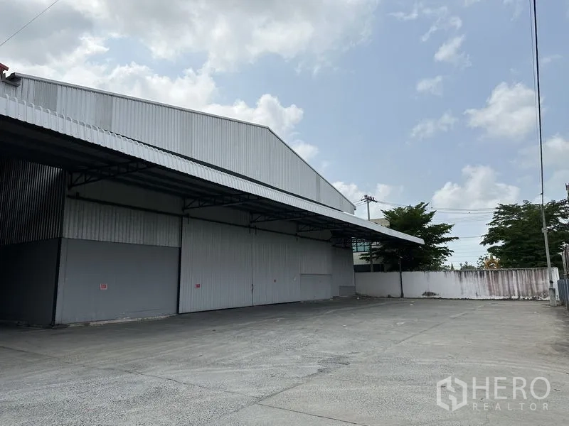 Warehouse for rent in Mueang Samut Prakan, Samut Prakan - Side exterior of the warehouse showing covered loading bay and a spacious concrete yard beside a perimeter wall and trees.
