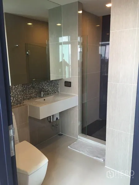 Condominium for rent in Khlong Toei, Bangkok - Contemporary bathroom featuring a glass-enclosed shower, wall-mounted sink, and neutral tiles.
