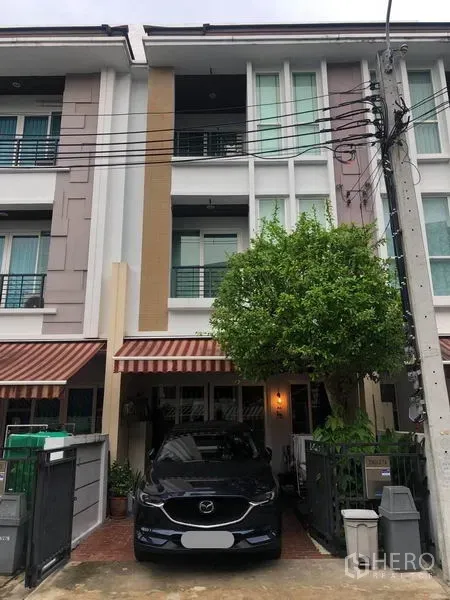 联排别墅 出售在Wang Thonglang, Bangkok - 三层联排别墅外立面，带车位、条纹遮阳篷和门前绿树，位于封闭社区内。