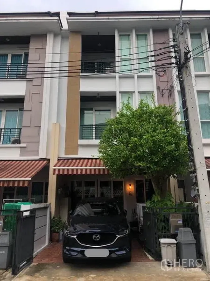 联排别墅 出售在Wang Thonglang, Bangkok - 三层联排别墅外立面，带车位、条纹遮阳篷和门前绿树，位于封闭社区内。