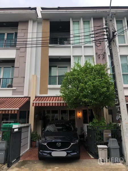 联排别墅 出售在Wang Thonglang, Bangkok - 三层联排别墅外立面，带车位、条纹遮阳篷和门前绿树，位于封闭社区内。