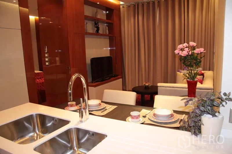 Condominium for rent in Khlong Toei, Bangkok - Open-plan kitchen with dining for two facing a cozy living area with TV and built-in shelving in a Khlong Toei condo.