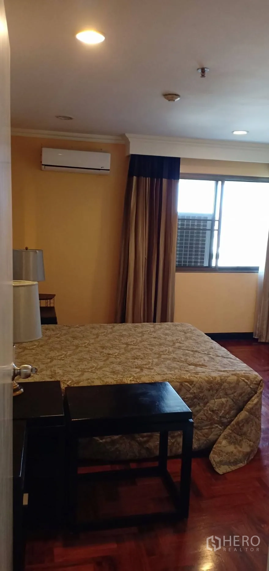 Condominium for rent in Watthana, Bangkok - Cozy bedroom with patterned bedding, bedside tables, a window with brown curtains and wall-mounted air conditioner.