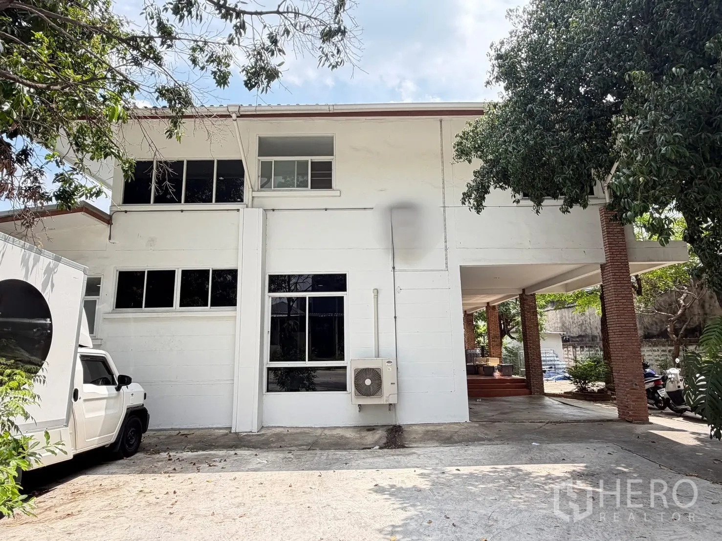 บ้านเดี่ยว for rent in Watthana, Bangkok - ภาพด้านหน้าบ้านเดี่ยวสองชั้นสีขาว มีเสาอิฐ โรงจอดรถ และต้นไม้ใหญ่ให้ร่มเงา