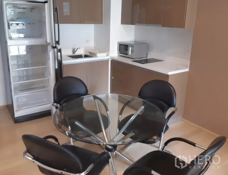 Condominium for rent in Khlong Toei, Bangkok - Modern kitchenette with glossy cabinets, sink, cooktop, microwave, tall fridge and a round glass dining table with four chairs.