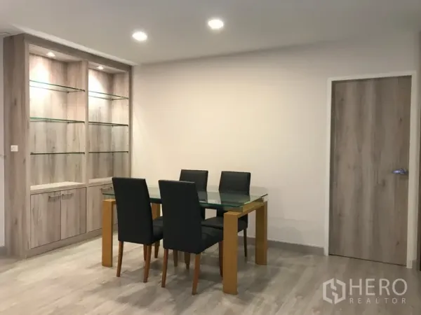 Condominium for rent in Watthana, Bangkok - Bright dining corner with a glass table, four black chairs, and illuminated glass‑shelf cabinet.