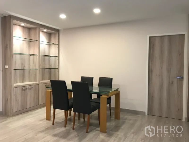 Condominium for rent in Watthana, Bangkok - Bright dining corner with a glass table, four black chairs, and illuminated glass‑shelf cabinet.