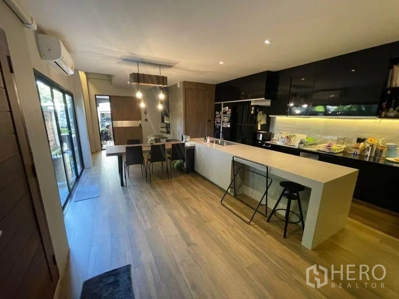 Townhouse for sale in Bang Phli, Samut Prakan - Open-plan kitchen with long island, black cabinets, pendant lights and wood flooring beside sliding glass doors.