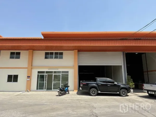 工厂 出售在Mueang Samut Prakan, Samut Prakan - 现代工厂外立面，橙色屋顶，玻璃办公区与大型卷闸门，外有皮卡停放。