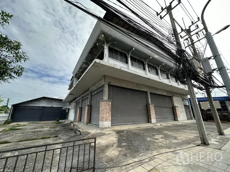 Home Office for rent in Khan Na Yao, Bangkok - Frontage with several metal roller shutters on a corner lot, sidewalk and side driveway leading to a rear warehouse gate.