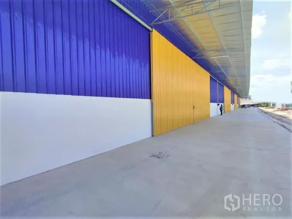 Warehouse for rent in Mueang Nonthaburi, Nonthaburi - Exterior walkway with a long canopy roof, multiple large sliding doors, and a broad concrete driveway.