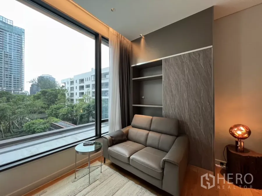 公寓 出租在Pathum Wan, Bangkok - 客厅配灰色皮沙发、嵌入式书架与落地窗，眺望绿树与城市景观。